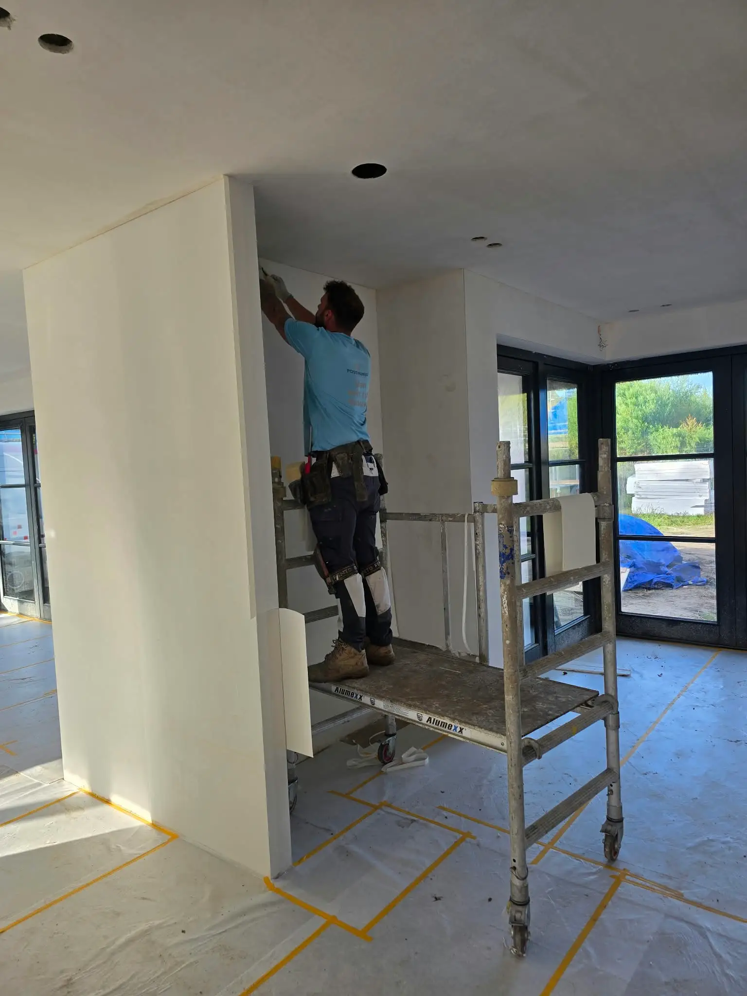 Schilder in bedrijfskleding van Posthumus Schilderwerken behangt vanaf een rolsteiger een wand in een nieuwbouwwoning